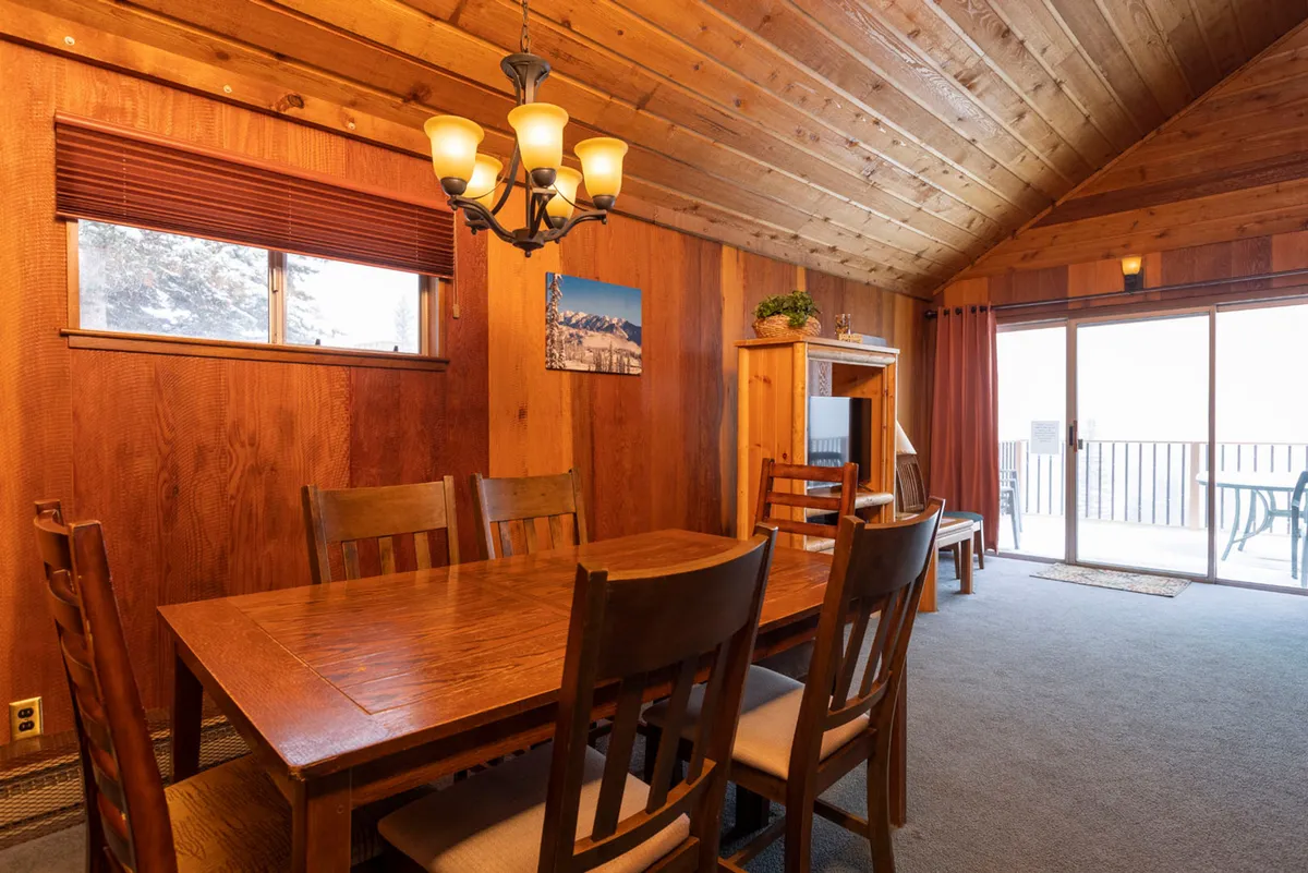 A dining area with a large wooden table, elegant chandelier, panoramic windows, and rustic wood-paneled walls. The space feels bright and open, perfect for group meals or board games.