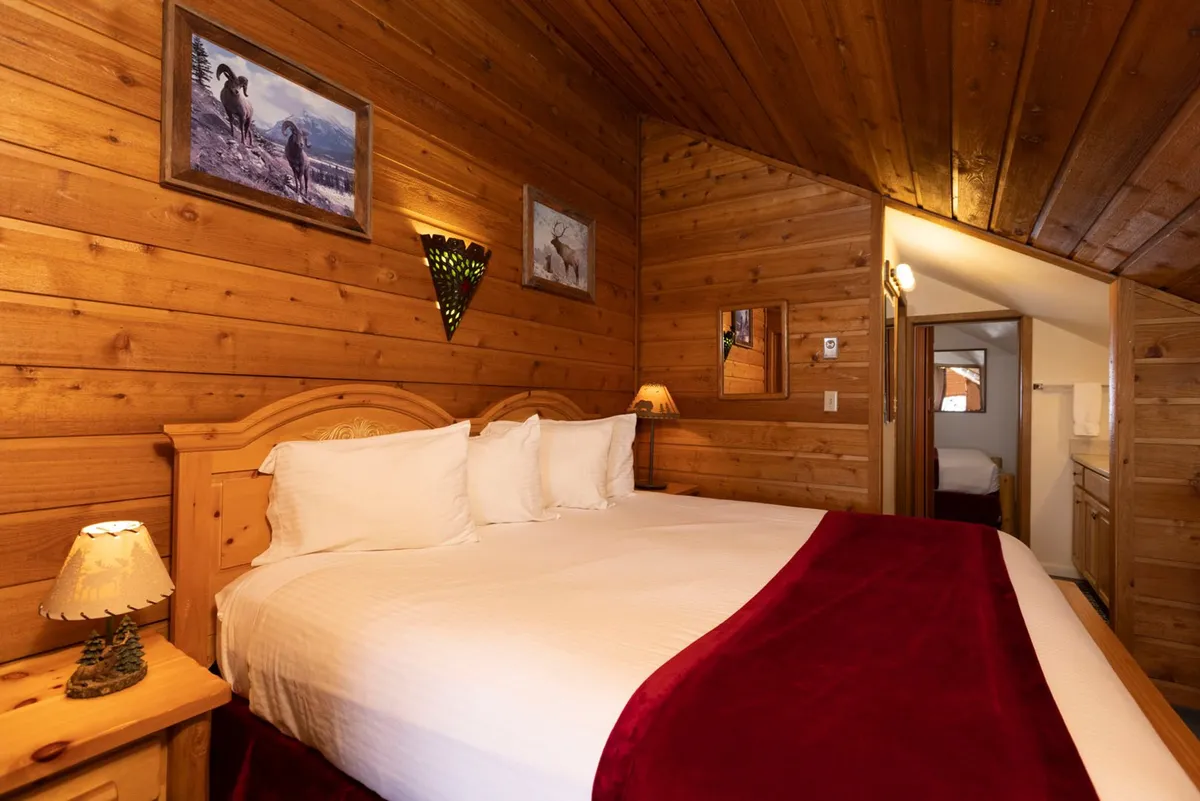A spacious, well-lit sleeping area featuring a large bed with white linens, a wooden headboard, and a decorative lamp. A piece of framed wildlife artwork adds a warm, personal touch. The scene feels both comfortable and open, ideal for a loft space.