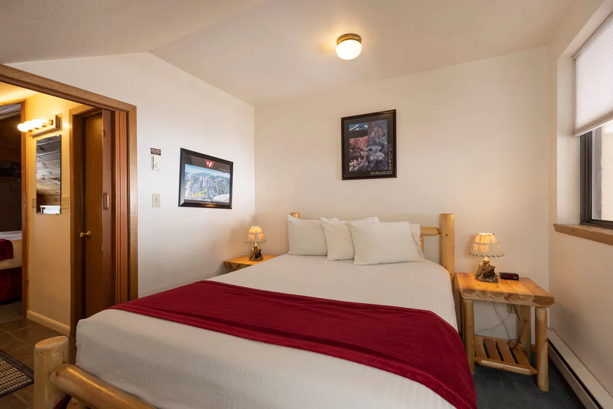 This image features a cozy bed adorned with a red throw blanket, flanked by wooden side tables and illuminated by table lamps. Decorative wall art adds a touch of personality, while the overall mood is relaxing and comfortable, inviting rest and quiet moments.