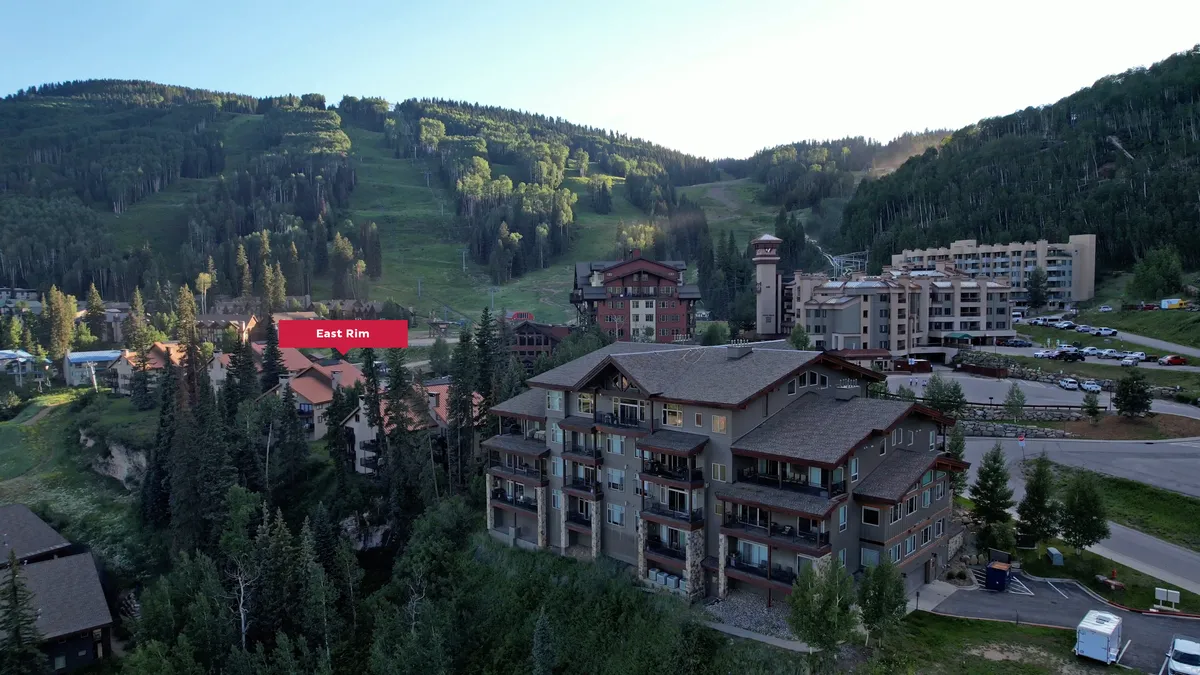 A tranquil exterior scene featuring residential buildings nestled against a dramatic mountain range, with wooded areas and a spacious parking zone. The image showcases the broader village context and easy access to mountain adventures.