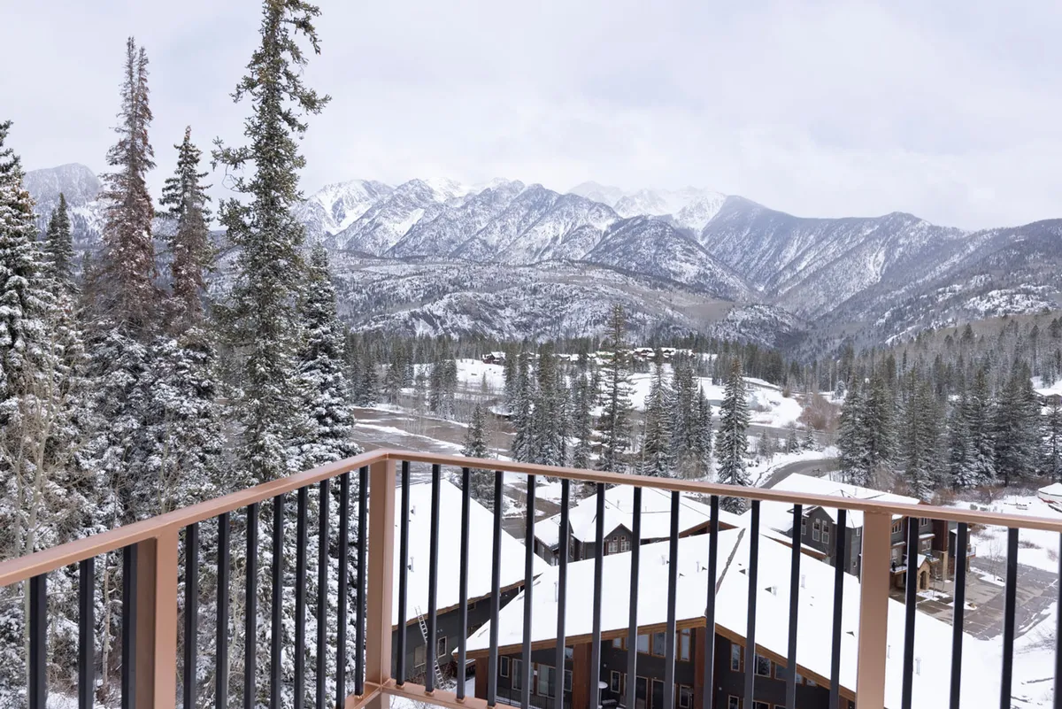 A tranquil view from a balcony overlooking snow-covered mountains, pine trees, and rooftops of nearby buildings. The scene hints at cozy relaxation after a day of exploration, with the balcony offering a private vantage point to soak in the scenery.