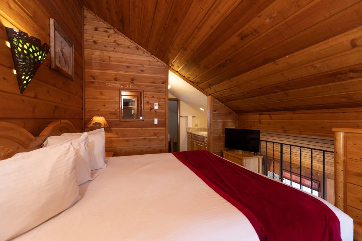 A warm, inviting bedroom setting with white linens accented by a red throw, wooden walls, decorative wall art, a small lamp, and a television. The space radiates coziness and a quiet retreat atmosphere, suitable for winding down after a long day.