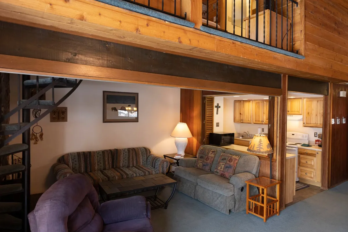 An interior scene showcasing a living area with a striped sofa (potentially convertible into a sleeper sofa), a spiral staircase, exposed wooden beams, and a glimpse of the kitchen. The space is inviting, emphasizing communal living and flexible use.