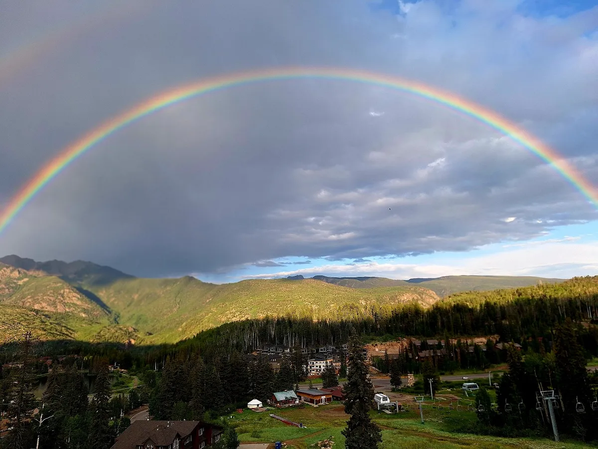 This hero image captures a breathtaking mountain landscape in Durango, Colorado, with a vibrant rainbow arching over a lush forest, a cozy alpine village nestled below, and a ski lift inviting adventure. The mood is uplifting and serene, perfectly showcasing experiences like hiking, skiing, nature walks, and capturing unforgettable photos.
