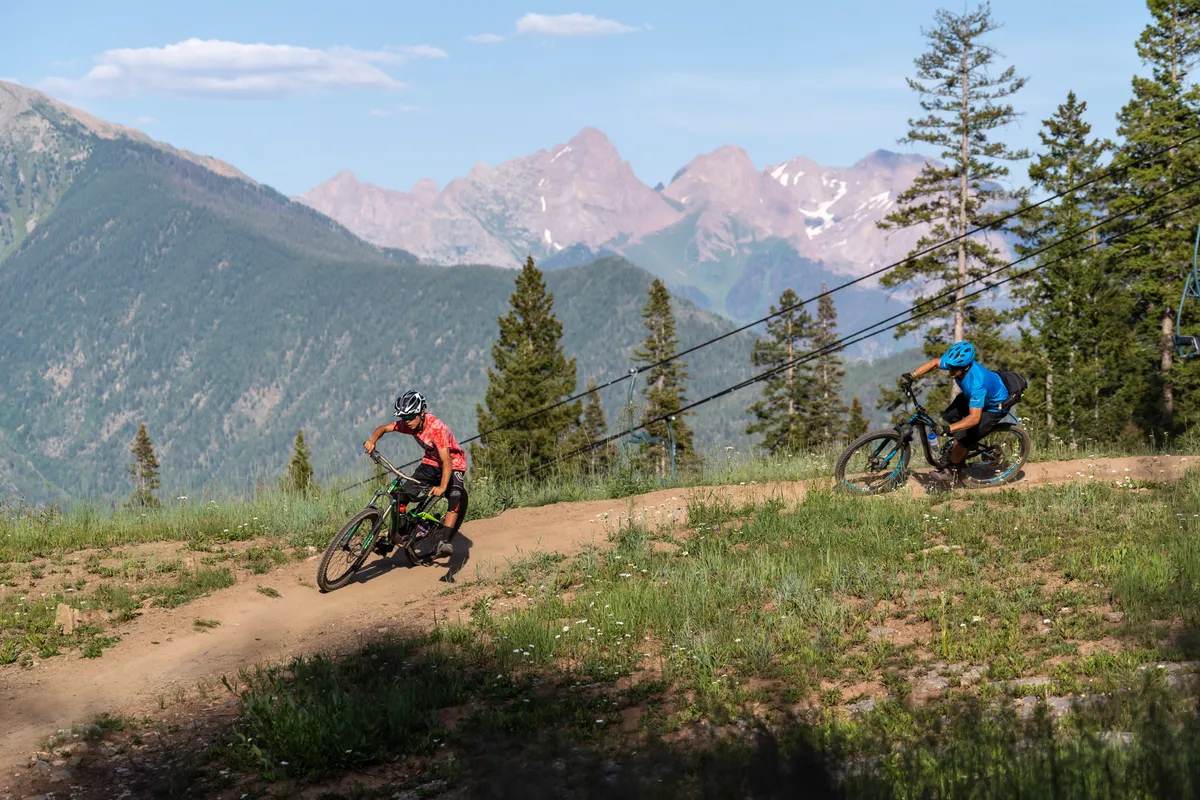 Mountain bikers navigate a rugged dirt trail framed by a dramatic mountain landscape. The scene is energetic and action-packed, emphasizing the thrill of outdoor activities available nearby. The sense of community among adventurers is palpable.