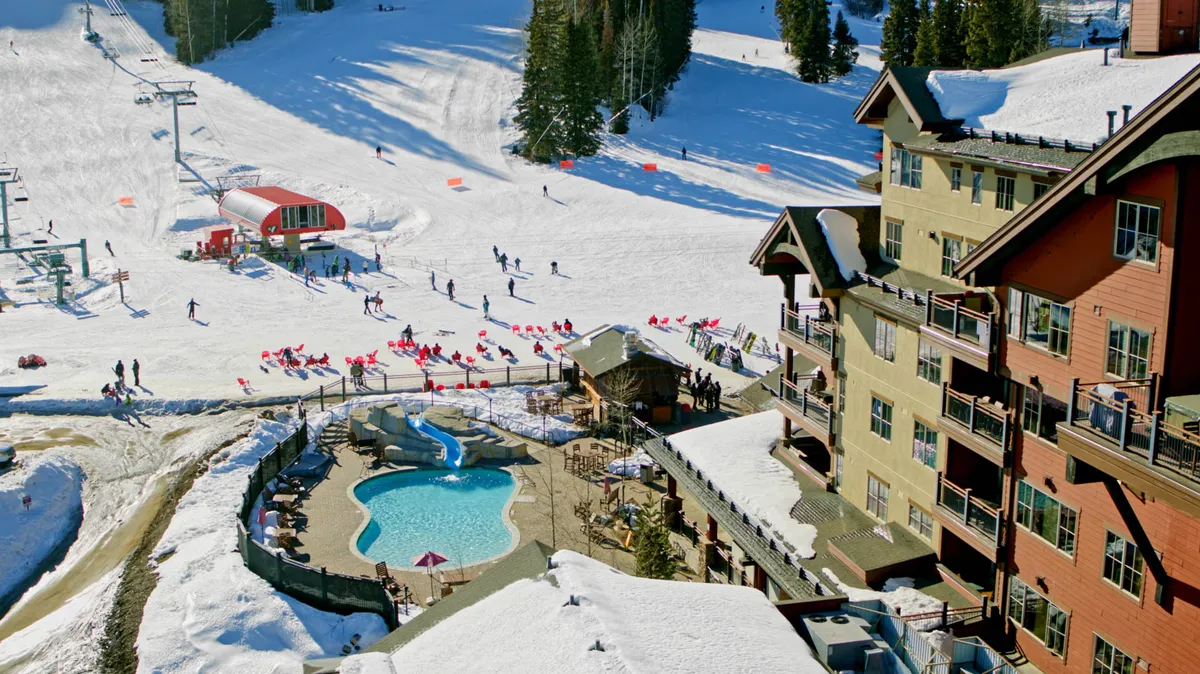 This vibrant exterior image displays the dynamic heart of the mountain village, showcasing bustling ski slopes, an inviting swimming pool, ski lift, and the lodge exterior backed by scenic mountain views. The scene is alive with energy, featuring guests participating in winter and après-ski activities.