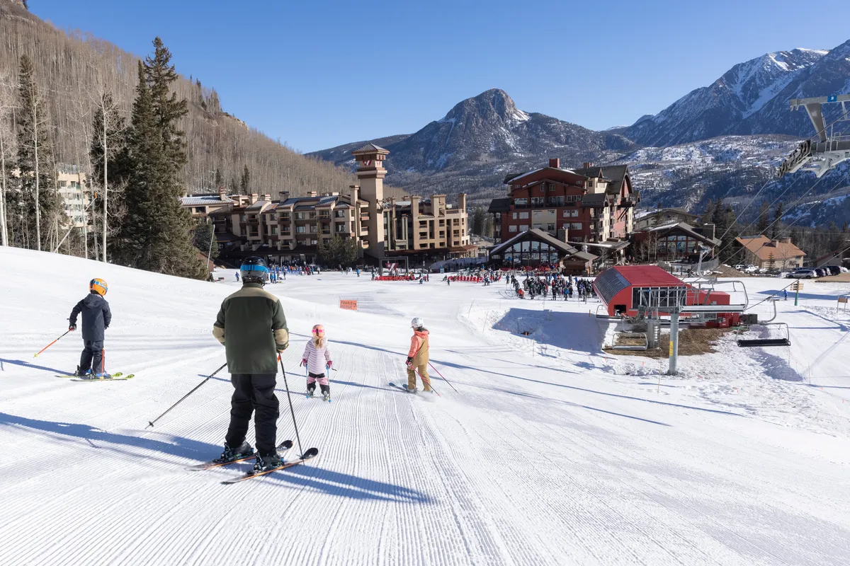 An exhilarating view of skiers descending a snowy slope, flanked by a majestic mountain backdrop and resort buildings. The scene buzzes with joyful activity, capturing families and groups making the most of the pristine winter environment. The visible ski lift and resort architecture reinforce the sense of community and accessibility.